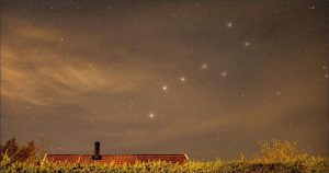 The Big Dipper constellation shines in the evening sky above a farmhouse and grassy landscape.
