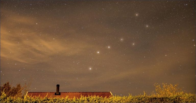 The Big Dipper constellation shines in the evening sky above a farmhouse and grassy landscape.