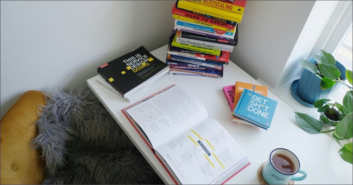 A desk with stacked books on work management methods, an open book, coffee and plant in window - Top work management methods