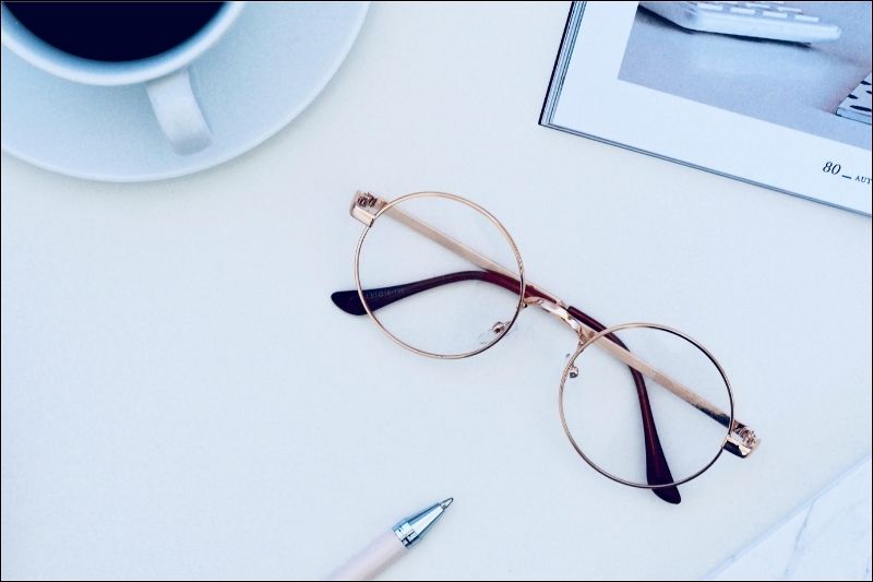 Grow Your Business With MKR Consulting Services Brown wire-frame glasses on a white surface with a coffee cup, pen, and the corner of a notebook.