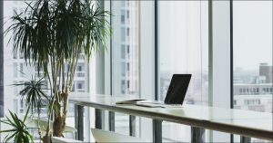 A laptop sits on a long table in front of a glass wall, overlooking a cityscape.
