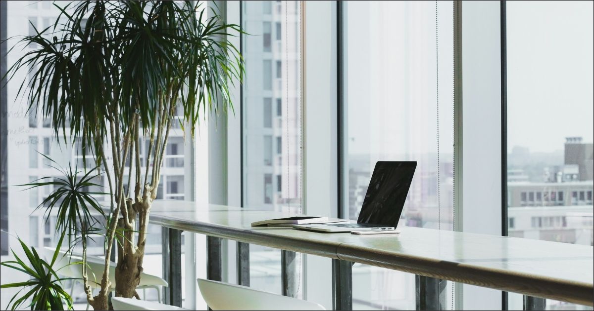 A laptop sits on a long table in front of a glass wall, overlooking a city scape.