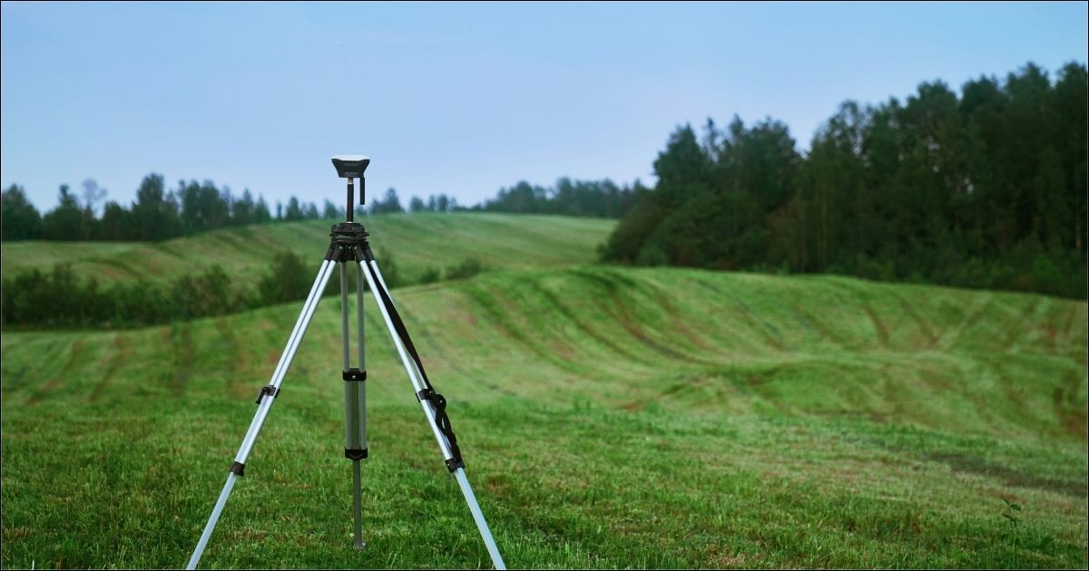 A survey scope set up in a green field, with a rolling landscape and tree lines.
