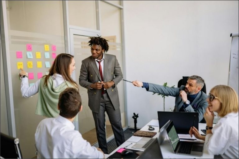A small work group collaborates around a table, using an ad-hoc Kanban board with sticky notes on a frosted glass wall.