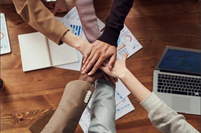 Hands of an office work team stacked together over a wooden table with papers and a laptop.